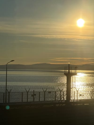 The media file depicts a sunrise over a body of water, likely a bay or sea, with land formations in the distance. A fence with barbed wire runs along the foreground, suggesting a secured area or perimeter. A lamppost is visible above the fence, its silhouette against the bright sky. The sun is low on the horizon, casting a golden glow on the water and illuminating thin clouds in the sky. The scene conveys a sense of calm and tranquility, with the warm colors of the sunrise creating an almost ethereal atmosphere. The location context points to Pevek, Russia, suggesting this scene captures an early morning in a coastal region. There is no visible text in the media file.