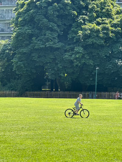 This media file features a person riding a bicycle on a well-maintained grassy field. The image is oriented sideways, with the top of the frame on the left and the bottom on the right.
The primary subject is a person, appearing to be a male, dressed in a light-colored t-shirt and shorts, riding a dark-colored bicycle. The bicycle has two wheels, handlebars, a frame, and pedals, and the person is actively pedaling.
The scene is an outdoor setting with a large, open expanse of bright green grass, indicative of a park or sports field. To the left of the grassy area, a dense canopy of green trees dominates the background, with sunlight filtering through the leaves. Beyond the trees, some indistinct buildings or structures can be seen. Along the edge of the grass field, there is a wooden fence. A tall, thin, dark pole with a light fixture stands near the fence, and some signs are also visible on the fence. In the distance, behind the fence, a few other people are seated.
The lighting suggests a bright, sunny day, likely mid-morning or afternoon, with strong shadows cast by the trees and the bicycle. The overall mood appears relaxed and recreational, given the activity and the pleasant outdoor environment.
There is no discernible text in the image. The location context provided is Molenbeek-Saint-Jean - Sint-Jans-Molenbeek, Belgium.