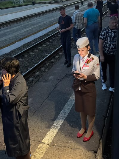 The image shows a group of people standing near railroad tracks. They are standing on an asphalt surface with painted white lines. The group includes a woman in a light gray uniform jacket, brown skirt, and red shoes, wearing a white peaked cap and the St. George ribbon. The woman is holding a small black object. Other people in the group are wearing casual clothing. The location is likely in Rtishchevo, Russia, given the person wearing the St. George ribbon.