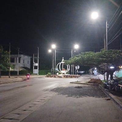 The image depicts the Crown Roundabout in Tanga, Tanzania, at night. The central feature is a large white sculpture composed of multiple concentric arcs, possibly representing a crown or stylized sphere, situated within a raised, brick-bordered circular island. Several bright streetlights illuminate the scene, casting light onto the asphalt road surfaces which show some irregularities. A single person on a red motorcycle, with its taillight illuminated, is visible on the left side of the road, moving towards the right. Utility poles with overhead power lines are present on both sides of the road and around the roundabout. To the far left, a multi-story white building with visible windows stands behind trees and a large, pole-mounted electrical transformer or similar utility structure. Green trees with dense foliage are positioned around the roundabout and along the road edges. A blue circular road sign indicating a roundabout and a black and white striped marker pole are visible on the central island and adjacent pavement, respectively.