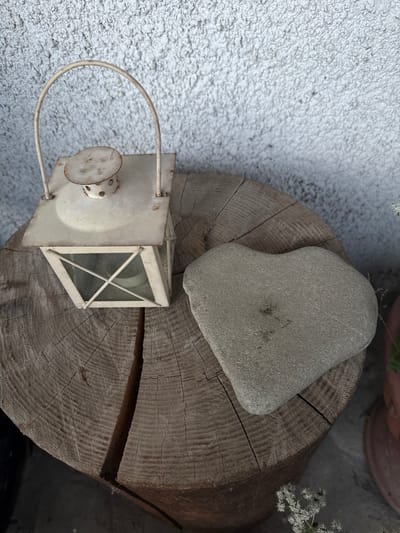 A still life composition is presented, featuring a vintage-style white lantern and a heart-shaped stone situated on a cross-section of a wooden log. The background is a textured, light-colored wall. The setting appears to be outdoors, possibly a courtyard or garden area in the city of Vyzhnytsia, Ukraine. The wooden log exhibits distinct growth rings and a deep crack running through it. The lantern is made of metal with glass panes and shows signs of age and wear. The heart-shaped stone is smooth and grey. The overall scene is static and devoid of any human presence or activity.
