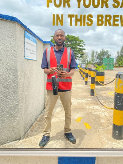 The image depicts a man standing in an outdoor industrial setting, possibly a brewery in Kinkinau, Nigeria, as suggested by the text overlay "IN THIS BREWERY". He is wearing a dark blue polo shirt, a red safety vest with reflective strips, and khaki pants. He is holding a black insulated water bottle in his left hand and a smartphone in his right hand. The man has a beard and is looking towards the camera. The background features a textured light-colored wall on the left, a row of black and yellow striped bollards with chains, and stacked green crates of beverages in the distance. The ground is paved concrete, with some yellow painted footmarks visible. The sky is overcast. The text overlay also includes "FOR YOUR SA" which is partially visible at the top of the image.