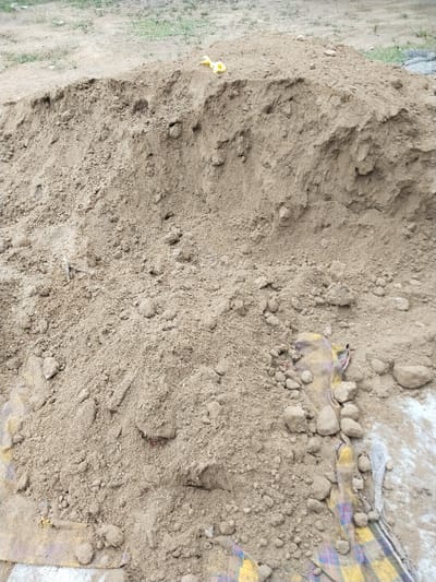 The image depicts a substantial, irregularly shaped mound of light brown, coarse-grained aggregate material, consistent with sand or sandy soil, located on unpaved ground in Jambo Dalam, Indonesia. The material exhibits varying particle sizes and includes scattered small stones. Two crumpled yellow plastic objects are visible on the highest point of the mound. At the lower right and left portions of the image, sections of a multicolored plaid fabric, featuring yellow, purple, and white patterns, are partially obscured by the aggregate material. The immediate surroundings consist of disturbed, barren earth with sparse patches of low-lying green vegetation in the background. No individuals or direct actions are visible within the frame.