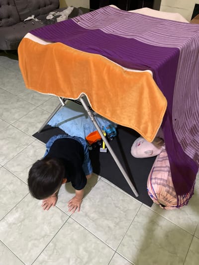 A child in a black shirt and blue shorts is crawling on a tiled floor. The child is near a structure covered with a purple blanket, an orange blanket, and a white pillow. Other items, including toys and a bolster pillow, are also near the structure. The scene is inside a room, likely in a residential setting. The location is Rojoimo, Indonesia.