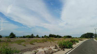 The image depicts a coastal scene with a road in the foreground, curving to the right. The road is paved and has a white dashed line marking its edge. To the left of the road is a sandy area with sparse vegetation, including bushes and some scattered rocks. In the midground, there is a line of trees and further back, the ocean is visible. The sky is partly cloudy with a mixture of blue sky and white clouds. The location is identified as Pusong, Indonesia. There are no people or animals visible in the image. There is a utility pole with wires on the right side of the road.
