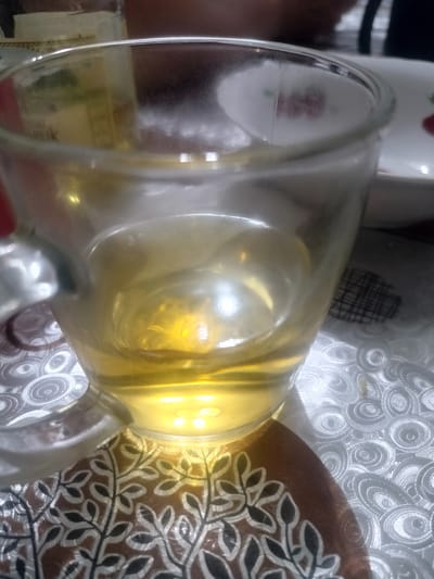 A close-up, eye-level shot captures a glass mug filled with a light-yellow liquid. The mug has a visible handle and is placed on a patterned surface. The surface exhibits two distinct patterns: one with swirling, circular designs and another with a floral motif of branches and leaves in dark and light tones. In the background, out-of-focus elements suggest a dining setting, with hints of a white plate adorned with a red floral design and what appears to be another beverage container. The lighting creates bright reflections on the liquid and the glass. The image is likely from Yarovoye, Russia, based on its context.