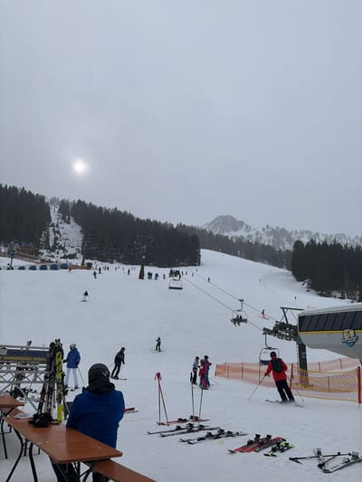 The image depicts an active ski resort on a cloudy winter day in Schwendau, Austria, characterized by a snow-covered mountain landscape under an overcast sky with a visible sun halo. The foreground features a snow-covered patio area with multiple wooden picnic tables and benches. One individual, wearing a blue jacket, is seated at a table with their back to the camera, observing the ski slope. Adjacent to the tables, several pairs of skis and ski poles are upright in a rack and laid flat on the snow.

In the midground, a wide, groomed ski slope descends from left to right. Numerous individuals, identified as skiers and snowboarders, are present on the slope. Some are actively skiing downhill, while others are static or conversing. A prominent chairlift system traverses the scene, with several chairs, some occupied by passengers, ascending the mountain. The lower station of the chairlift, partially visible on the right, features a building with a "Mayrhofner" logo. A skier in a red jacket is navigating towards this station. Orange safety netting delineates areas on the slope near the chairlift.

Further up the mountain on the left, a multi-story building with a brown roof and blue railings is visible, potentially a restaurant or ski school, indicated by a partially legible sign "SPORT SCHIEST... SNOWBOARDSCHULE RAMSAU." Dense evergreen forests cover the mountainous terrain, particularly on the left and extending across the background. Snow-dusted mountain peaks are visible in the far background. Light snowfall appears to be occurring.