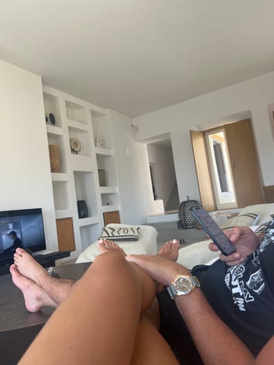 The image depicts an indoor scene within a modern, minimalist living space, consistent with properties found in Agios Ioannis Diakoftis, Greece. Two individuals are present, observed from a low, first-person perspective, likely from the lap of one individual looking towards the other.
In the immediate foreground, a pair of bare, tanned legs, presumably belonging to a female (based on painted toenails with light polish), are crossed and resting on a dark, flat surface, possibly a coffee table. Beyond these legs, on the right, the lower body and arm of a male individual are visible. He wears a black t-shirt with white graphics, partially showing "199". His right forearm, adorned with a gold-toned wristwatch featuring a light grey or green fabric strap, is holding a dark-colored smartphone vertically with both hands, thumb positioned on the screen. His bare feet are also visible, resting on a light-colored, textured sofa or lounge chair.
The room is brightly lit with natural light, featuring white walls and a white ceiling. On the left side, a large built-in shelving unit with multiple recessed rectangular niches displays various decorative objects: dark abstract sculptures, a circular light-colored object on a stand, white abstract sculptures, a large light-brown textured vase, and a dark vase. The bottom two niches contain rectangular woven baskets. To the left of this shelving unit, a flat-screen television is visible, displaying an image of a person in a dark suit.
In the background, on the right, a light-colored wooden door or shutter is partially open, revealing a bright outdoor space. Next to this, a large, dark, woven lantern or decorative object sits on the floor. A central archway or doorway leads to a hallway with stairs descending into a lower level of the dwelling. The flooring throughout the visible area appears to be light-colored. The overall environment suggests a contemporary, tranquil, and comfortable setting.