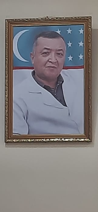A framed portrait hangs centrally on a light-colored wall in Kogon City, Uzbekistan. The portrait depicts a middle-aged man of Central Asian ethnicity, with short, dark hair, a round face, and a subtle smile. He is wearing a white lab coat over a dark, collared shirt. The background of the portrait features the national flag of Uzbekistan, characterized by a light blue upper stripe containing a white crescent moon and multiple white stars, followed by a thin white stripe and a thin red stripe. The portrait is enclosed within an ornate, gold-colored rectangular frame, which is suspended from the wall by a visible hanging mechanism at the top. The wall itself is a uniform, plain surface.