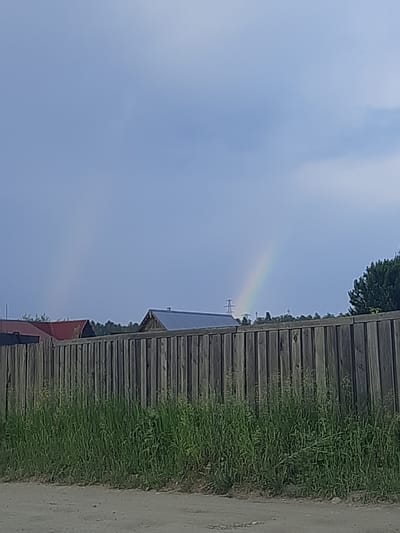 The image captures an outdoor scene with a wooden fence in the foreground. Behind the fence, several buildings with red and metallic roofs are visible, suggesting a rural or suburban setting. Tall green grass grows in front of the fence. In the sky, a faint rainbow arc can be seen, indicating recent or ongoing precipitation. A power line tower is also visible in the distance. The overall atmosphere suggests a moderately cloudy day. The location is Shelekhov, Russia.