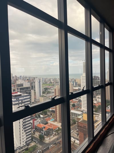 The image presents an elevated, wide-angle view of a dense urban cityscape from a building interior, looking out through a large, multi-paned window. The window frame, a dark, matte material, divides the view into a grid-like pattern. The cityscape is characterized by a variety of modern and older buildings, ranging from tall high-rises with distinct architectural features, such as the white building with blue vertical accents and the building with a dark grey facade, to lower-rise residential structures with terracotta roofs. Several buildings appear to be under construction, evidenced by visible cranes and scaffolding.

The urban environment extends towards a body of water, likely the Atlantic Ocean, which forms the horizon under a cloudy sky. The sea appears calm and stretches across the width of the image. The sky itself is overcast with grey and white clouds, with hints of brighter sky peeking through in some sections, suggesting diffused daylight.

The foreground includes the interior of the room from which the photograph is taken, with a dark, possibly wooden, surface visible along the bottom right edge. The perspective suggests the viewer is on a high floor of a building in João Pessoa, Brazil, a coastal city known for its numerous skyscrapers and proximity to the ocean. The buildings' architectural styles and density are consistent with urban development in a Brazilian coastal metropolis. The street level is visible in some parts of the image, showing roads with parked and moving vehicles, indicating active city life. The overall impression is one of a sprawling, active urban center situated by the sea.
