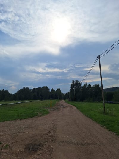 The image shows a dirt road winding through a rural landscape. On the left side of the road, there is a green grassy field with a long white pipe running through it. A yellow marker post is visible in the field. A line of trees forms a dark green border on the horizon behind the field. On the right side of the road, a wooden utility pole with two wires stretches across the frame. Beyond the pole, more trees and a grassy area are visible. The sky is partly cloudy with the sun breaking through, casting a bright light. The overall impression is one of a rural, undeveloped area, possibly in the city of Atamanovka, Russia.