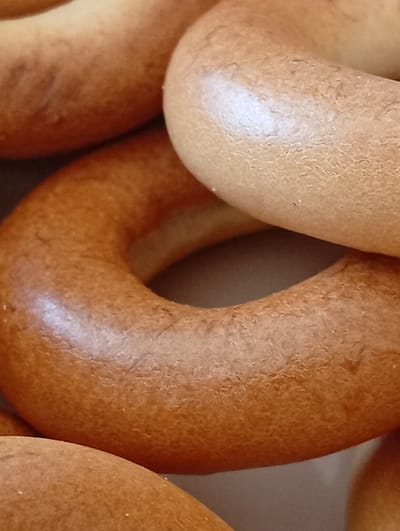 A close-up, overhead shot displays a pile of golden-brown, ring-shaped bread products, possibly bagels or kalatchi. The bread has a slightly textured surface with visible imperfections and a glossy sheen in some areas, indicating a baked finish. The objects are arranged in a way that suggests a stack or collection, with the focus on the round shapes and warm hues. The background is indistinct and appears to be a neutral surface, allowing the bread to be the primary subject. There are no people or discernible elements of a city environment, including the city of Buțeni, Moldova. The image is purely a still life of baked goods.