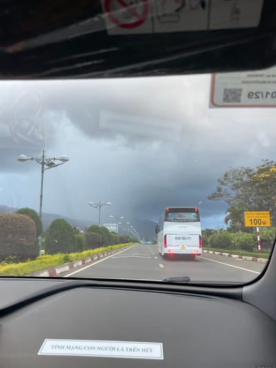 The image shows a view from inside a vehicle, looking out at a highway scene in Liên Nghĩa, Vietnam. The sky is overcast with dark clouds, suggesting impending rain or a storm. A white tour bus with the license plate "50H-361-77" is traveling on the right side of the highway. On the right side of the road, a yellow road sign indicates "100M" with Vietnamese text above it. Lush green bushes and trees line the roadside, with ornamental yellow flowers visible in the distance. Lampposts are spaced along the highway. In the foreground, on the dashboard of the vehicle, there is a white sticker with green text that reads "TÍNH MẠNG CON NGƯỜI LÀ TRÊN HẾT", which translates to "Human life is above all". The overall impression is a view of a highway journey under a dramatic sky.