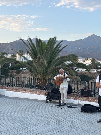 A musician, an elderly man with white hair and fair skin, plays an acoustic guitar on a paved outdoor terrace. He is dressed in a white outfit, including pants and a long-sleeved shirt. He holds the guitar across his lap, with his right hand strumming and his left hand on the fretboard. A microphone is positioned in front of him.

Behind the musician, a large palm tree dominates the left side of the frame, its fronds reaching across the scene. Beyond the palm tree and the black metal railing of the terrace, a picturesque view unfolds. White buildings with terracotta roofs are nestled on a hillside, overlooking a valley. In the distance, rugged mountains rise against a pale blue sky dotted with wispy white clouds.

To the right of the musician, a black portable speaker with the "JOYO" logo is visible, along with a black backpack and some cables. The terrace is paved with light-colored tiles arranged in a diamond pattern, and a white ledge runs along the edge. The lighting suggests it is late afternoon or early evening, with a soft glow illuminating the scene. The overall atmosphere is serene and relaxed, with the musician likely performing for an audience or simply enjoying the moment. The location cues, such as the architecture and the Mediterranean vegetation, strongly suggest the setting is Nerja, Spain.