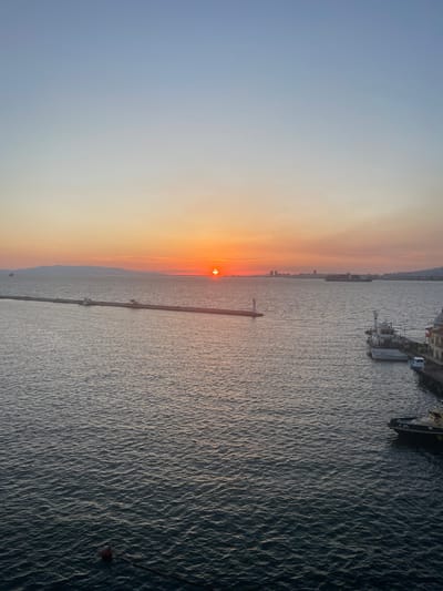 The image depicts a serene sunset over the Aegean Sea in İzmir, Turkey. The sun, a vibrant orange disc, is low on the horizon, casting a warm glow across the sky. The sea is calm, with gentle ripples reflecting the sunset's colors. In the foreground, a red buoy floats on the water, attached to a dark rope.

A long, concrete pier stretches out into the sea, punctuated by a small lighthouse and a couple of structures. Further out, a large container ship, the CMA CGM, sails in the distance. Along the right edge of the image, a portion of a dock is visible, with several boats moored alongside, including a white tugboat. In the far distance, the İzmir skyline is silhouetted against the colorful sky. The overall atmosphere is peaceful and tranquil, capturing the beauty of a coastal sunset.