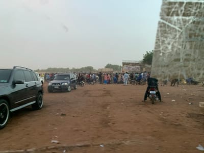 The scene takes place in Sokoto, Nigeria at latitude 13.049 and longitude 5.238. The image was captured from a car, looking out the window. There are approximately 50 people, all dressed in casual attire, gathered in an open dirt lot. Some are standing and talking, others are sitting on motorcycles and cars. A man in a dark shirt and pants is riding a motorcycle towards the camera. There are several cars and motorcycles parked in the lot. The day is overcast and dusty. The lot appears to be located in a residential or commercial area. A building with a large billboard can be seen in the distance. The billboard is plastered with colorful posters and writing. Trees are visible in the background.
