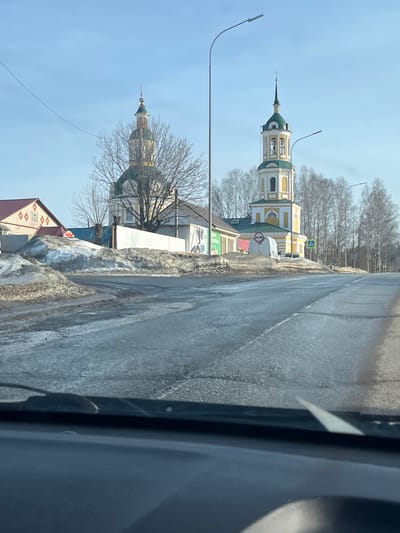 The image depicts a static street scene in Чутырь, Russia, captured from the interior of a vehicle, evidenced by the visible dashboard and the lower edge of the windshield. The environment suggests late winter or early spring conditions, with a clear blue sky and bright sunlight.
In the mid-ground, two distinct religious structures, characteristic of Russian Orthodox architecture, are prominent. To the left, a church building features a central golden dome surmounted by a green onion dome and a cross, positioned above a larger green dome; its walls are light in color. To the right, a multi-tiered bell tower stands, predominantly yellow and white with green roof sections and a green spire topped by a golden finial and cross. Bare deciduous trees are interspersed in the background and around these structures.
The foreground displays an asphalt road with visible cracks, patches, and a somewhat wet or icy appearance. Large, dirty snowdrifts line the shoulders of the road. Two streetlights with curved poles extend over the roadway. To the far left, a building with a reddish roof and decorative gables is partially visible behind snow. Between the church and bell tower, low-rise commercial buildings with various signage are partially obscured by a white wall or fence. Road signage includes a circular red 'No Entry' or 'No Stopping' sign and a blue square 'Pedestrian Crossing' sign, both situated near the bell tower. A partially visible white van is positioned behind the red sign. No individuals are present in the visible scene.