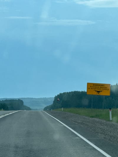 The image shows a view of a two-lane asphalt road stretching into the distance, with a lush green forest on either side. The sky is overcast with a light blue hue. On the right side of the road, a yellow rectangular sign with black lettering is prominently displayed. The sign has the text "ВНИМАНИЕ! Контроль выезда на встречную полосу!" (ATTENTION! Control of driving into the oncoming lane!) and features a black silhouette of a drone at the bottom. There are also two white road markers with red tops visible along the right edge of the road. In the distance, rolling hills covered in trees are visible. The overall scene depicts a rural highway in Karagay, Russia, with a warning sign about traffic control.