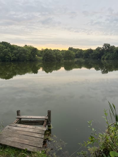 A serene lake scene unfolds with a weathered wooden pier in the foreground, its planks showing signs of age and decay. The pier juts out into the still water, which reflects the overcast sky and a line of lush green trees bordering the far bank. The trees are dense and varied, creating a natural, undisturbed landscape. A faint glow of sunset or sunrise can be seen on the horizon, painting the sky with soft hues of yellow and orange. The water is calm, with only a few ripples disturbing its glassy surface. The overall impression is one of tranquility and natural beauty. The location is the city of Istobnoye, Russia.