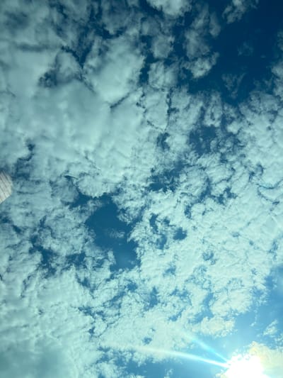 The image displays a bright blue sky filled with scattered, fluffy white clouds. A sun flare, emitting bright white and blue light rays, is visible in the lower right corner of the frame. A portion of a striped object, possibly part of a flag or banner, is present in the upper left quadrant. The scene is set in Sânnicoară, Romania. There are no people or specific objects indicative of urban activity; the focus is entirely on the sky and atmospheric conditions. The overall impression is a clear, sunny day with cloud formations.