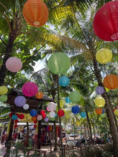 This image, taken in Tân Thạch, Vietnam, depicts a vibrant outdoor market scene framed by palm trees adorned with colorful paper lanterns. The lanterns are suspended at varying heights and span a wide range of colors including red, orange, yellow, green, blue, purple, and white. The market area is lined with stalls displaying various items, some of which appear to be clothing, textiles, and other wares. People can be seen browsing and interacting with the vendors. The image is taken from a low angle, looking upwards towards the lanterns and the sky visible through the palm leaves.