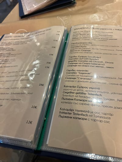 The image shows a page from a menu in Limenaria, Greece, located at the coordinates 40.628, 24.575. The page features a list of seafood dishes written in Greek and German. The menu is printed on white paper with a blue cover. Some of the dishes include: Garides Saganaki (Shrimp Saganaki), Kalamari Sxaras yeimosto (Stuffed Calamari), and Frittierter Tintenfisch mit Tartarsauce (Fried Squid with Tartar Sauce). The prices are listed in Euros. The page is open to reveal the next page, which is also part of the menu. The menu is laying on a table, likely in a restaurant. The image does not show any people.
