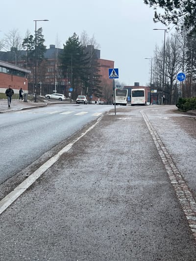 This image depicts an overcast daytime scene on a wet urban street in Espoo, Finland, specifically near Vitikka 1, 02630, adjacent to the Espoo Courthouse. The ground, including the asphalt road and paved pedestrian/cycle paths, appears damp, possibly from rain or melting ice/snow.

The foreground features a two-lane asphalt road on the left, curving towards the right in the mid-ground. A white solid line delineates the left edge of the road. To the right of the road is a wide, dark-paved pedestrian and cycle path, distinguished by a double line of reddish cobblestones running along its right edge. Both surfaces are speckled with dark grit.

On the left side of the road, a male figure in a dark jacket and trousers walks away from the camera. Further down the left sidewalk, another individual in a white top and dark trousers is partially visible. A white station wagon is parked uphill on the left, facing away from the camera, with a dark SUV behind it.

In the mid-ground, two white articulated city buses are stopped at a bus station on the right side of the road, both facing away from the camera. The rear of one bus clearly displays "oike." Behind the buses, a bus shelter with a yellow roof and blue base is visible. A pedestrian in dark clothing stands near the bus stop on the far right.

Prominently placed near the center of the image is a blue rectangular sign indicating a pedestrian crossing. Further right, a blue circular sign with white bicycle and pedestrian symbols indicates a shared path. Multiple dark-poled streetlights illuminate the area. In the background, several multi-story reddish-brown brick buildings with dark roofs are visible, interspersed with bare deciduous trees and dark green conifer trees.
