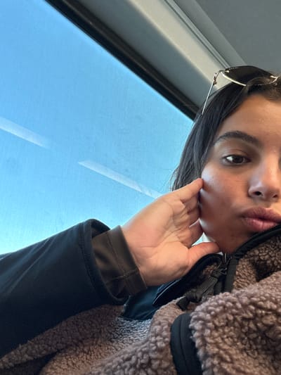 A female individual is depicted in a selfie-style image taken inside a vehicle, located in Secaucus, United States. Her right eye is visible, and her lips are slightly pursed. Dark hair is partially visible. Her right hand is positioned with fingers curled against the right side of her face, with the thumb resting near the jawline. She wears a black long-sleeved garment beneath a dark brown or grey sherpa-style fleece jacket with a visible zipper and black trim. Dark-lensed sunglasses, bearing a "Gucci" logo, are perched on top of her head. A large window occupies the upper left portion of the frame, revealing a clear blue sky. The window exhibits horizontal streaks and reflections, suggesting motion. Dark window framing and a light-colored interior panel are visible above the window.