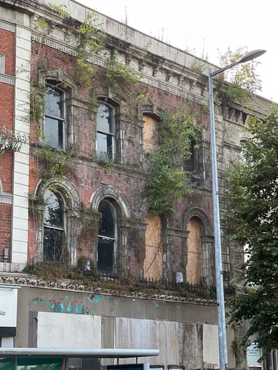 This is a photograph of an abandoned building located on Great James Street in Derry/Londonderry, United Kingdom. The building, constructed of red brick, exhibits significant decay and overgrowth. Green vines and plants climb the facade, partially obscuring windows and architectural details. Some windows are covered with wooden boards, while others have mesh coverings. The upper portion of the building features decorative stonework and cornices, also intertwined with vegetation. A street light stands in front of the building, to the right of the frame. Below the upper floors, the lower section of the building shows peeling paint in shades of green, red, and beige, with more boarded-up windows. The overall impression is one of neglect and the reclaiming of urban space by nature.