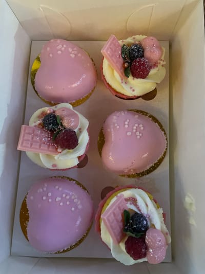 In Kobeliaky, Ukraine, a white cardboard box contains six heart-shaped desserts. Three desserts are on the left side of the box, arranged vertically, and three are on the right side, also arranged vertically. All desserts have a pink frosting or glaze. The top left dessert is a heart shape with white dots on its pink surface. The middle left dessert is a cupcake with white frosting, topped with a pink chocolate bar, raspberries, blackberries, and pink sprinkles. The bottom left dessert is a heart shape with pink frosting and white dots. The top right dessert is a cupcake with white frosting, adorned with a pink chocolate bar, raspberries, blackberries, pink candies, and pink sprinkles. The middle right dessert is a heart shape with pink frosting and white dots arranged in a "16" pattern. The bottom right dessert is a heart shape with pink frosting and white dots.