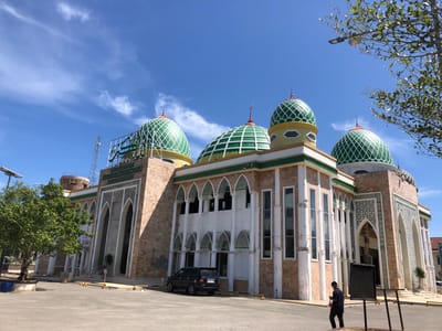 The image shows the Masjid Agung Darunnayim mosque in Gampongjawa, Indonesia. The mosque features a sand-colored facade with white pillars, green domes with white latticework, and arched windows and entrances. A sign in Arabic script sits atop one section. A black SUV is parked in front of the building. A man in dark clothing walks along the pavement in front of the mosque. The sky is blue with scattered clouds. A tree is visible on the left side of the image. Streetlights and other infrastructure elements are present.