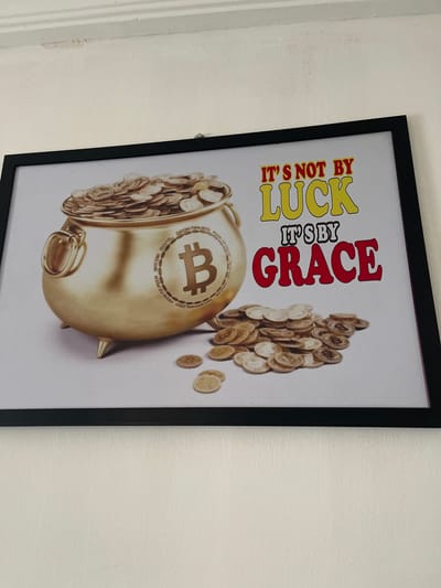A framed print hangs on a white wall. The print features a gold pot overflowing with gold coins bearing the Bitcoin symbol. More Bitcoin coins are scattered next to the pot. Above the pot, text reads "IT'S NOT BY LUCK" in yellow and orange, and "IT'S BY GRACE" in red. The location, Naze, Nigeria, is not detectable from the given visual information.