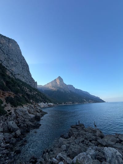 A scenic view of the coastline near Baunei, Italy, features rugged, rocky shores leading into the calm, deep blue sea. Jagged cliffs rise steeply on the left, covered in sparse green vegetation, and a prominent, pointed mountain peak is visible in the distance, catching the light of the setting sun. Three people are standing on the rocks in the foreground, two of them near the water's edge and one further back, appearing to be observing the view or fishing. The sky is a clear, pale blue, indicating it is either early morning or late evening.