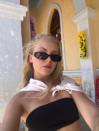 A young woman with blonde hair and fair skin is captured in a close-up, sideways shot. She is wearing black sunglasses with gold detailing on the sides and a black halter top. A light-colored, patterned scarf is tied around her neck, its ends draped over her shoulders. Her lips are slightly parted, and her expression appears relaxed.

The background suggests an outdoor setting with architectural elements. A yellow wall with decorative trim is visible, adorned with a garland of bright yellow flowers. To the left, a portion of an arched window with stained glass can be seen. Below this, a row of delicate pink flowers sits on a ledge. The overall impression is of a warm, possibly sunny day. The lower portion of the image shows a textured, light-colored surface, likely a wall or pavement. The scene is situated in Hội Tín, Vietnam, which aligns with the floral decorations and architectural style. No specific activity is taking place; it appears to be a posed portrait. No discernible text is present in the image.