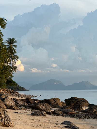 A sandy beach in Baan Pang Ka, Thailand is dotted with dark, jagged rocks of various sizes. To the left, lush green trees and palm trees line the edge of the beach, their fronds reaching towards the sky. The sea stretches out to the horizon, calm and serene, with hazy mountains visible in the distance. The sky above is filled with dramatic, billowing clouds in shades of blue and white, with some areas illuminated by soft, golden light, suggesting either sunrise or sunset. The overall scene is one of natural beauty and tranquility.