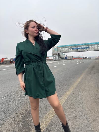 A young woman stands outdoors on a paved road in what appears to be an industrial or transportation area in Minderevo, Russia. She is positioned slightly to the left of the frame, facing partially right and looking upwards with a neutral expression. Her dark hair is long and appears windswept, framing her face. She is wearing a dark green, short-sleeved dress or tunic with a tie-waist and dark boots. A dark red watch is visible on her left wrist, and a light-colored bracelet on her right. Her left hand is raised towards her head, while her right hand lightly holds the hem of her dress.

The ground is grey asphalt or concrete, marked with white dashed lines and a solid yellow line, indicating a road or large paved surface. In the background, to the right of the woman, a prominent blue-framed structure resembles a covered pedestrian walkway or bridge. Clearly visible on this structure is the Cyrillic text "РОСАТОМ" (ROSATOM), which is the Russian State Atomic Energy Corporation, providing a significant location cue. Further back, vehicles, including what appears to be a large tanker truck, and indistinct buildings can be seen. The sky is uniformly overcast and light grey, suggesting a cloudy day.