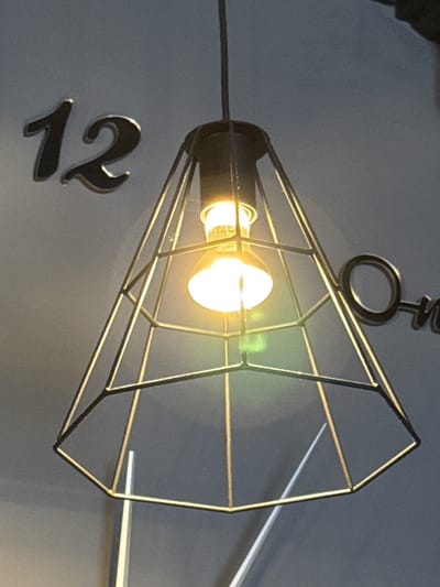 The image shows a close-up of a modern pendant lamp with a geometric metal cage. The lamp is lit, with a bright filament visible inside. Behind the lamp, the numbers "12" are visible, appearing to be metallic or reflective numbers affixed to a wall. To the right of the lamp, partially obscured by the metal cage, is the word "one" also appearing to be metallic or reflective. The background is a plain, light gray wall. The scene suggests an indoor environment, possibly a cafe or a modern living space. The location in Kirishi, Russia is not discernible from the visual information provided.