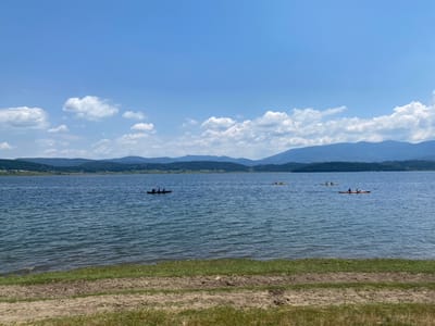 This image depicts a scenic lake scene, likely taken at Tsigov Chark, Bulgaria. The photograph is oriented upside down, with the sky and clouds appearing at the bottom and the lake and shoreline at the top.
In the water, several individuals are engaged in water activities, with what appear to be kayaks or canoes visible. One kayak, with two occupants, is positioned in the middle-left of the frame. Another appears to be a single-person kayak to its right, and further right, a larger boat with multiple people is visible. The water is a calm blue, reflecting the sky above.
The shoreline is a strip of land with grass and dirt, suggesting a natural bank. Beyond the lake, rolling hills and mountains form a picturesque backdrop. A band of fluffy white clouds stretches across the horizon, separating the lake from the clear blue sky above.
The lighting suggests it is daytime, likely a sunny day with pleasant weather conditions, suitable for outdoor water recreation. The overall mood is peaceful and recreational, showcasing a beautiful natural landscape and people enjoying their time outdoors. No visible text is present in the image.