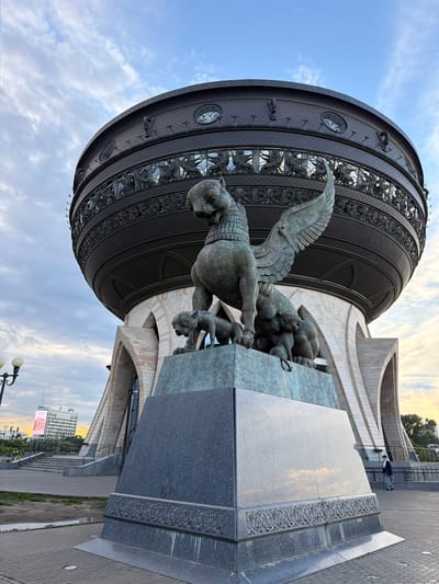 The image shows a large, spherical building with intricate metallic ornamentation and a bronze sculpture in the foreground. The sculpture depicts a winged lion-like creature grappling with smaller creatures. The building features a textured, dark metallic facade with decorative circular elements and what appear to be relief sculptures of mythical beings. The architectural style is modern, with curved white stone elements contrasting with the dark facade. A paved walkway and grassy areas are visible in the background, along with a modern building in the distance and streetlights. The sky is partly cloudy with a warm glow suggesting either sunrise or sunset. The overall scene evokes a sense of grandeur and artistry. There is a person standing on a walkway in the middle distance, appearing small in comparison to the monumental sculpture and building. There is also a large digital billboard visible in the distance with some text and images on it. The image appears to be taken outdoors during daylight hours, possibly during the golden hour. The location is Shelanga, Russia, as indicated by the context.