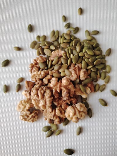 A pile of walnuts and pumpkin seeds are scattered on a white textured surface. The walnuts are broken into pieces, revealing their natural brown color and textured interior. The pumpkin seeds are a vibrant green and oval-shaped. Some seeds are clustered with the walnuts, while others are spread out individually across the surface. The background is a white, subtly patterned material, possibly a placemat or cloth. There is no indication of people, actions, or interactions. The scene is set on a flat surface, with no specific environmental context suggesting a location in Кривец, Russia.