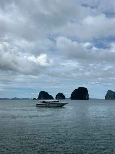 An outdoor, eye-level shot captures a serene seascape in Tambon Krasom, Thailand. The ocean, with its subtle ripples, occupies the majority of the frame, reflecting the overcast sky. Two speedboats navigate the water; one is closer to the foreground on the left, while another is further out in the center. The boats are white with blue accents and appear to be carrying passengers. In the distance, several large, green, rocky islands rise dramatically from the sea, their forms softened by the hazy atmosphere. The sky is a mosaic of grey and white clouds, with occasional patches of blue peeking through. The overall impression is one of tranquil natural beauty and maritime activity.