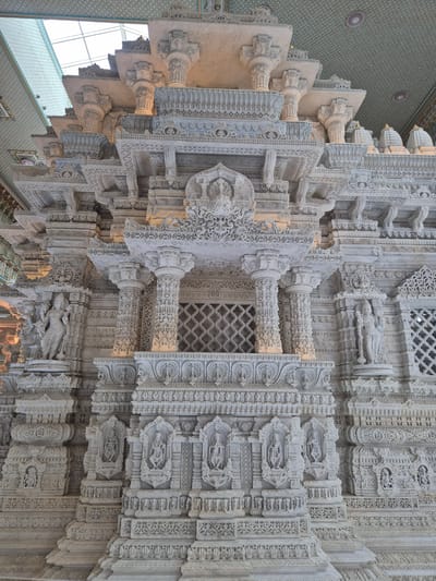 This is a photograph of the exterior of the BAPS Swaminarayan Akshardham temple in Robbinsville Township, New Jersey, United States of America. The temple is constructed of white marble and features intricate carvings and detailed sculptures. The image focuses on the ornate facade of the structure, showcasing multiple levels of carved balconies, pillars, and decorative elements. Statues of deities and figures are visible within niches and on pedestals. The architecture is highly detailed and exhibits traditional Indian temple design. The environment appears to be indoors, as suggested by the presence of a large skylight and a patterned ceiling. There are no people visible in the image.