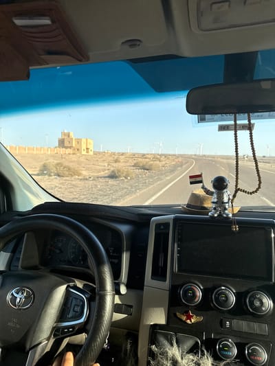 The view from inside a vehicle moving along a road with a roadside building on the left, in the Gharib Cape area of Egypt, is captured. The steering wheel, dashboard, and rear-view mirror are visible in the vehicle's interior. A rosary and an astronaut figurine holding an Egyptian flag hang from the rear-view mirror. The controls on the dashboard feature buttons and an attached star pin. The speedometer shows the date and time, and the Toyota logo is visible on the steering wheel. The exterior shows an arid landscape, with buildings and vegetation. Wind turbines are visible in the distance.