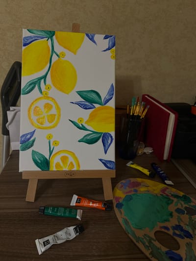 Here is a detailed description of the image:
The image shows a still life of an artist's setup. In the foreground is a canvas propped on a small easel. The canvas has a painting of lemons and leaves on it, done in yellow, green, and blue hues. Behind the canvas, various items are arranged, including a red notebook, a jar of paintbrushes, a small container with shells, tubes of paint (yellow, blue, orange, green, and white), and a wooden palette covered in blobs of dried paint.
The canvas is situated at the bottom of the frame. The other art supplies are above and slightly behind it. The background consists of a light brown, textured surface. The overall setting appears to be indoors.