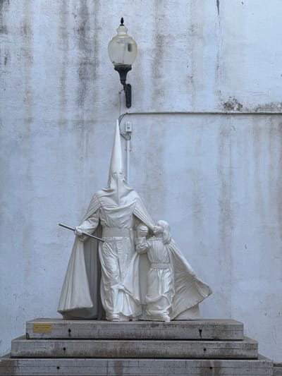 The image shows a statue of the Virgin Mary carrying a figure in the attire of a penitent, likely a Nazareno, as evidenced by the pointed capirote hood. The statue is white and appears to be made of stone or plaster. It's mounted on a concrete structure attached to a textured white wall. A streetlight with a glass globe extends from the wall to the left of the statue. The background wall shows signs of weathering and staining. A small plaque is affixed to the concrete structure beneath the statue. The statue is located in Vinaròs, Spain.
