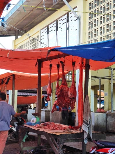 This image captures a vibrant open-air meat market stall in Tuha Gp Gajah, Indonesia, under bright, diffused daylight. Orange and blue tarpaulin canopies provide shade over the scene. In the mid-ground, a wooden table serves as a display for raw meat, covered with brown paper and laden with piles of red meat cuts, offal, and bones. A green mechanical weighing scale with a circular pan is positioned on the table to the left. Directly above this stall, five large cuts of raw meat, predominantly reddish-pink hindquarters with hooves visible, are suspended by metal hooks from a horizontal beam. A larger, irregularly shaped mass of raw meat hangs centrally among them.
To the left, a man viewed from behind, dressed in a light blue short-sleeved shirt and reddish-brown pants, stands near a black and white scooter. His right arm is slightly raised. Partially obscured by the hanging meat are the lower bodies of at least two other individuals, one wearing dark pants and boots, the other shorts. A striped, light-colored towel hangs from a wooden support pole on the right side of the stall, with a red fabric strip also hanging vertically nearby.
The background features a multi-story yellow building characterized by rectangular ventilation blocks and horizontal windows. Beyond the immediate stall, additional orange canopies stretch into the distance, covering more market areas with visible corrugated metal roofs. A red scooter is partially visible on the far right. The ground beneath the stall is uneven and shows scattered debris. Overhead, multiple ropes and wires crisscross, supporting the canopy structures.