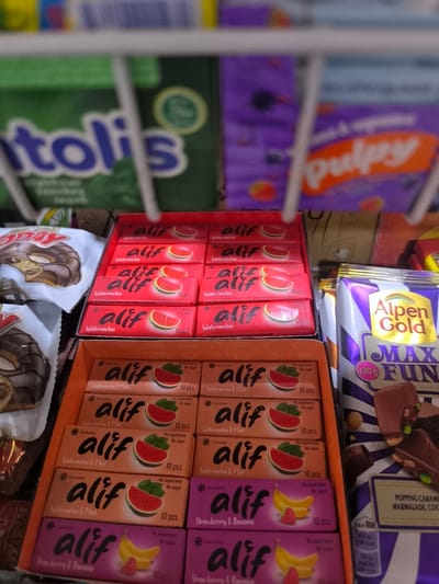 The image shows a close-up of a display of packaged chewing gum and chocolate bars in a store. In the foreground, several rows of "Alif" brand chewing gum are visible, with packaging featuring images of watermelon slices and bananas, and text indicating "Watermelon," "Watermelon & Mint," and "Strawberry & Banana" flavors. Some packs also state "No aspartame" and "No sugar." Behind the gum, a large bar of "Alpen Gold" chocolate, labeled "MAX FUN" and weighing 130g, is displayed. Further in the background, blurred images of other products, including green packaging with the word "itolis" and purple packaging with the word "pulpsy," are visible. The products are arranged on shelves or in a display case. The lighting suggests an indoor setting, likely a retail environment. There are no people or actions depicted in the image. The location in Yangikishlak, Uzbekistan, is inferred from external context and is not directly observable in the image itself.