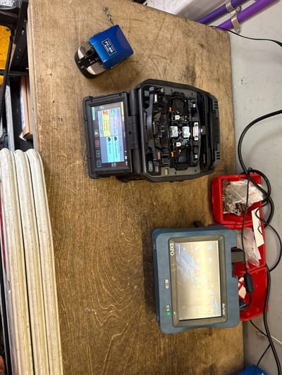 The image shows a close-up, overhead view of a wooden workbench. The workbench is cluttered with various tools and equipment related to fiber optic splicing.
In the top left quadrant of the image, a blue fiber optic cleaver is visible. To its right, a large, open fiber optic splicing machine is positioned. The machine has its internal components exposed, revealing delicate machinery and wires. Its screen displays text and graphics related to fiber optic splicing parameters.
Below the splicing machine, a red plastic container holds small metal parts and tools. A black cable snakes from the splicing machine towards the right side of the image, connecting to an unseen power source.
In the bottom right quadrant, a blue handheld device, identified by the "EXFO" logo as a MaxTester, is prominent. Its screen displays a touch interface with various icons and menus, suggesting it's in an active state, possibly performing diagnostics or settings configuration. A green indicator light is visible on the front of the device. Another black cable connects to this device, similar to the one connected to the splicing machine.
To the left of the workbench, a stack of white, coiled material, likely fiber optic cable spools, is partially visible. The background of the image shows a light-colored wall and some purple pipes secured with metal brackets.
The lighting in the scene is adequate, casting soft shadows and highlighting the details of the equipment. The overall impression is that of a workspace actively being used for fiber optic installation or maintenance.
The scene described is not located in the city of Watford, United Kingdom.