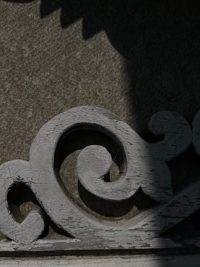 This is a close-up, low-angle shot of weathered, white-painted wooden scrollwork against a textured, gray stone background. The scrollwork features a prominent spiral design. Sunlight casts sharp shadows across the scene, highlighting the peeling paint and rough texture of the wood and stone. The image is taken outdoors, with a hint of dappled light and shadow suggesting natural elements or an architectural setting. There are no people or animals visible. The overall impression is one of aged beauty and the passage of time. It's impossible to determine the exact location in Preobragenskaya, Russia, from the visual information provided.